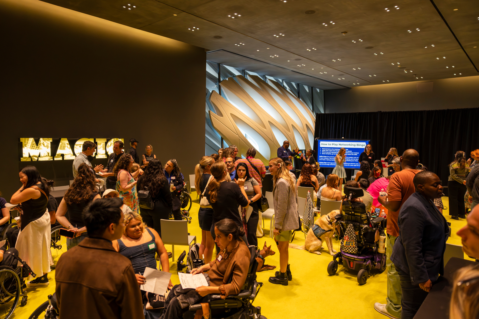 A packed room at The Broad with an illuminated MAGIC installation, attendees in wheelchairs and standing, networking on a bright yellow floor.